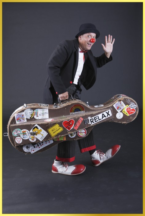 Der Musikclown tmit roter Pappnase und Cellokoffer in der Hand, winkt in die Kamera..